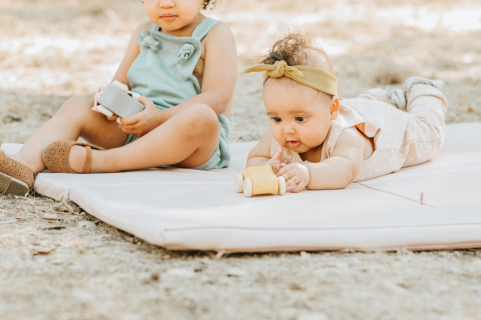 a baby and a toddler playing with toys outside on their toki mats play mat